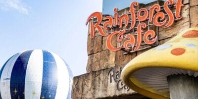 Rainforest Cafe sign set against a rugged rock wall, with colorful themed decor including a striped hot air balloon and an oversized yellow mushroom creating an immersive, adventure-filled atmosphere.