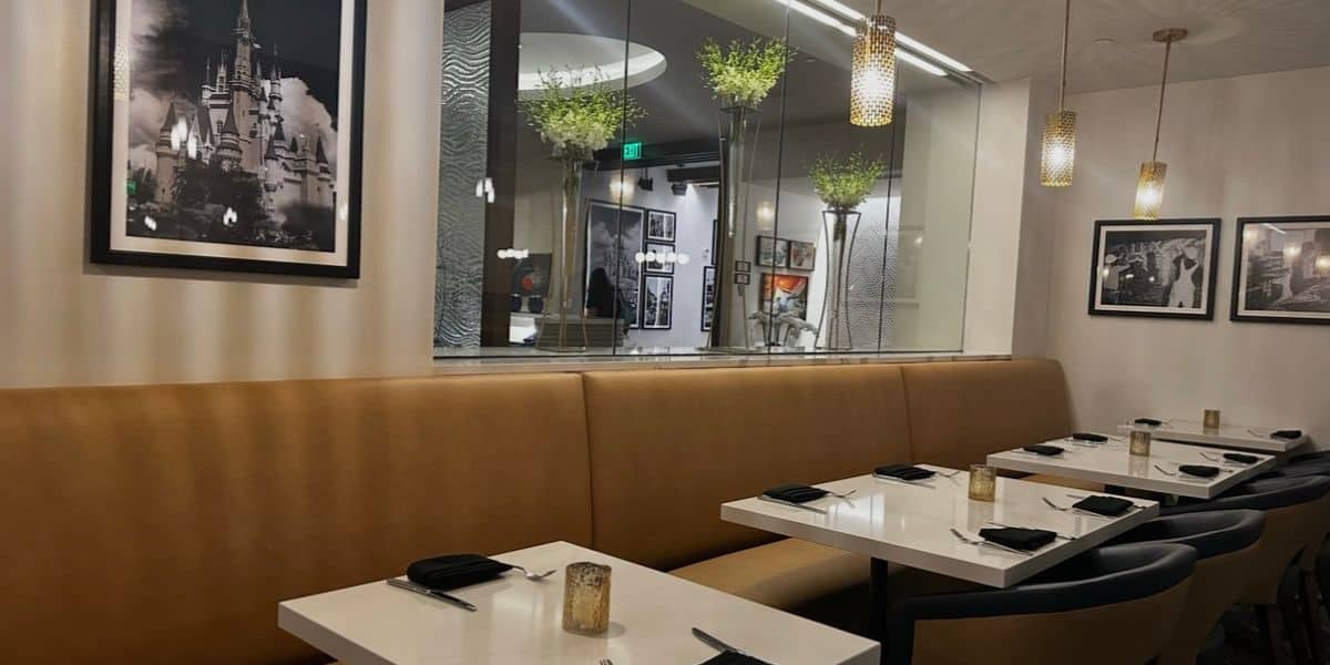 Vibrant dining area with comfy tan booths, crisp white tables set with black napkins, playful wall art, and a glass partition filled with lush greenery—perfect for families fueling up before hitting the park’s thrilling attractions.