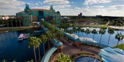 The Swan and Dolphin Resort features towering swan statues, lush palm trees, and a scenic walkway spanning over sparkling water, all set against a backdrop of partly cloudy skies—an inviting oasis steps away from the Walt Disney World magic.