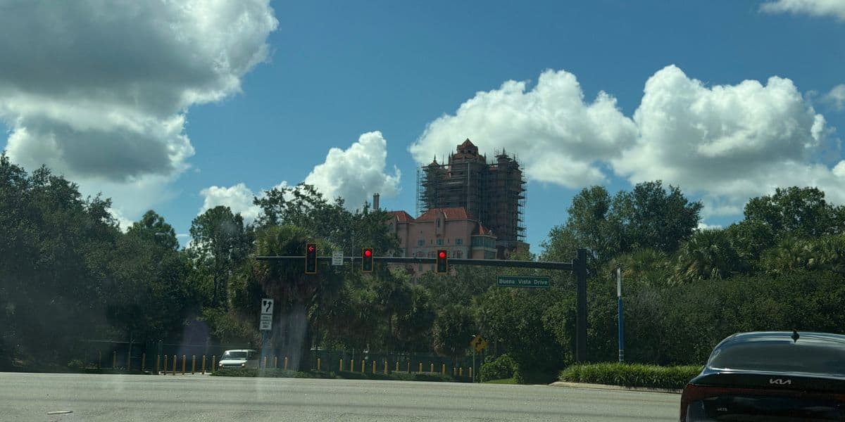 A vibrant pink theme park attraction undergoing renovations stands behind lush greenery, viewed from the main park walkway under a bright blue sky filled with whimsical clouds.