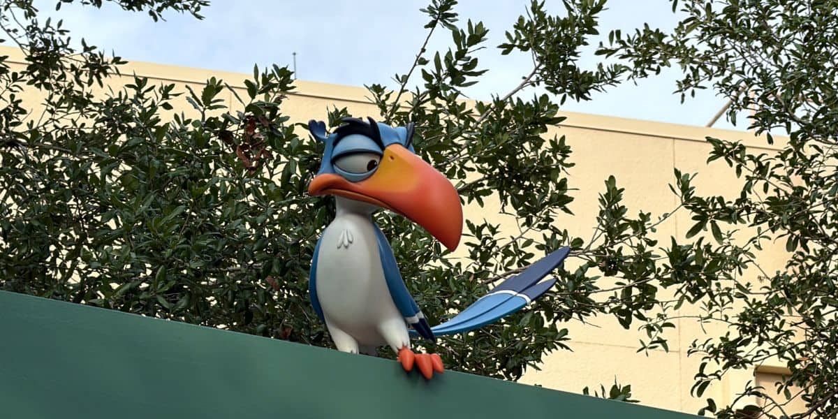 A vibrant, animated bird with an oversized beak perches on a green ledge, set against a backdrop of lush trees and an iconic park building, capturing the playful spirit of beloved Disney characters found throughout Hollywood Studios.