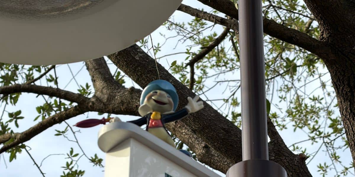 Jiminy Cricket, channeling classic Disney magic, greets guests with a cheerful wave from atop a white sign nestled beside leafy green trees, bringing New Attraction excitement to the park.