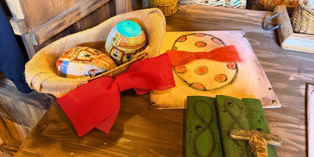 Two toy Easter eggs nestled in a burlap basket with a large red bow and a festive drawing on a wooden table.