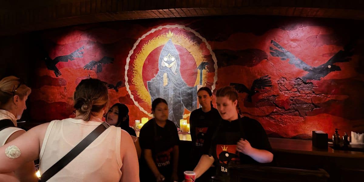 Staff serving customers at Oga’s Cantina in Star Wars: Galaxy’s Edge in front of a large mural featuring Six Ravens and a hooded owl.