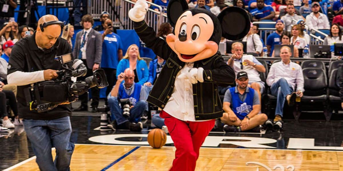 Mickey Mouse waving on the Orlando Magic basketball court at Disney World with a cameraman nearby.